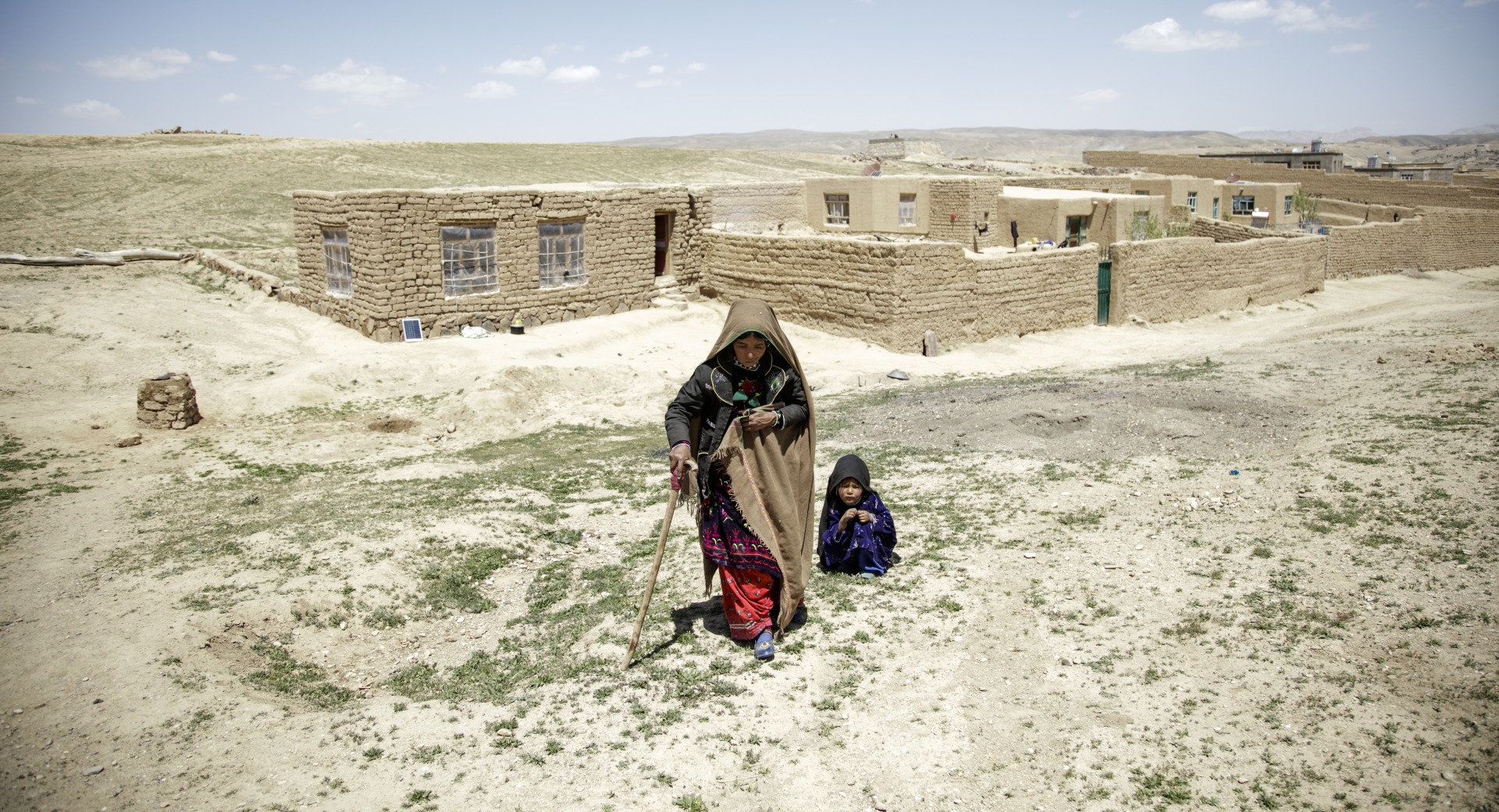 A mother walks away from her home with the help of a cane. Her daughter crouches behind her.