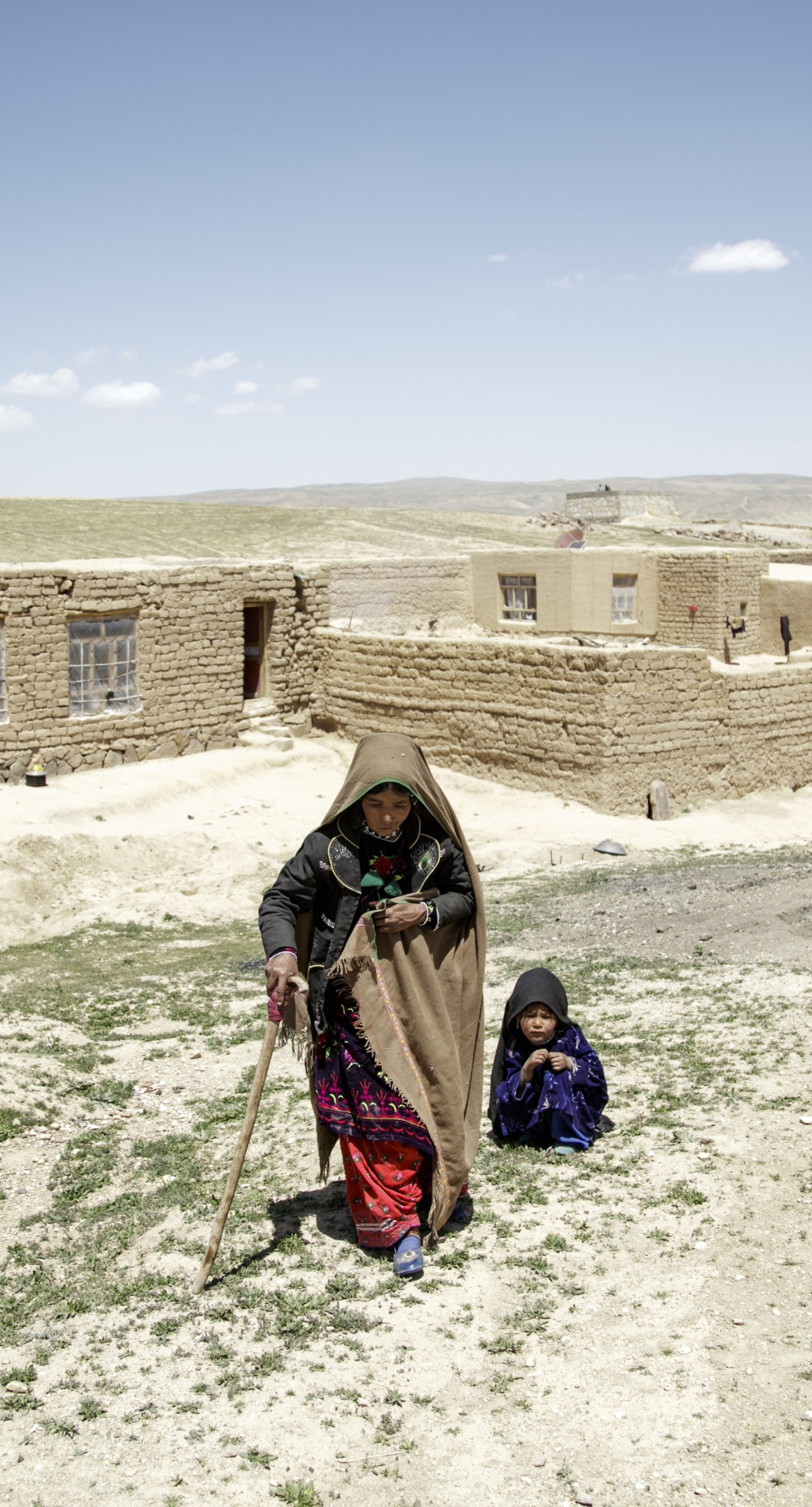 A mother walks away from her home with the help of a cane. Her daughter crouches behind her.