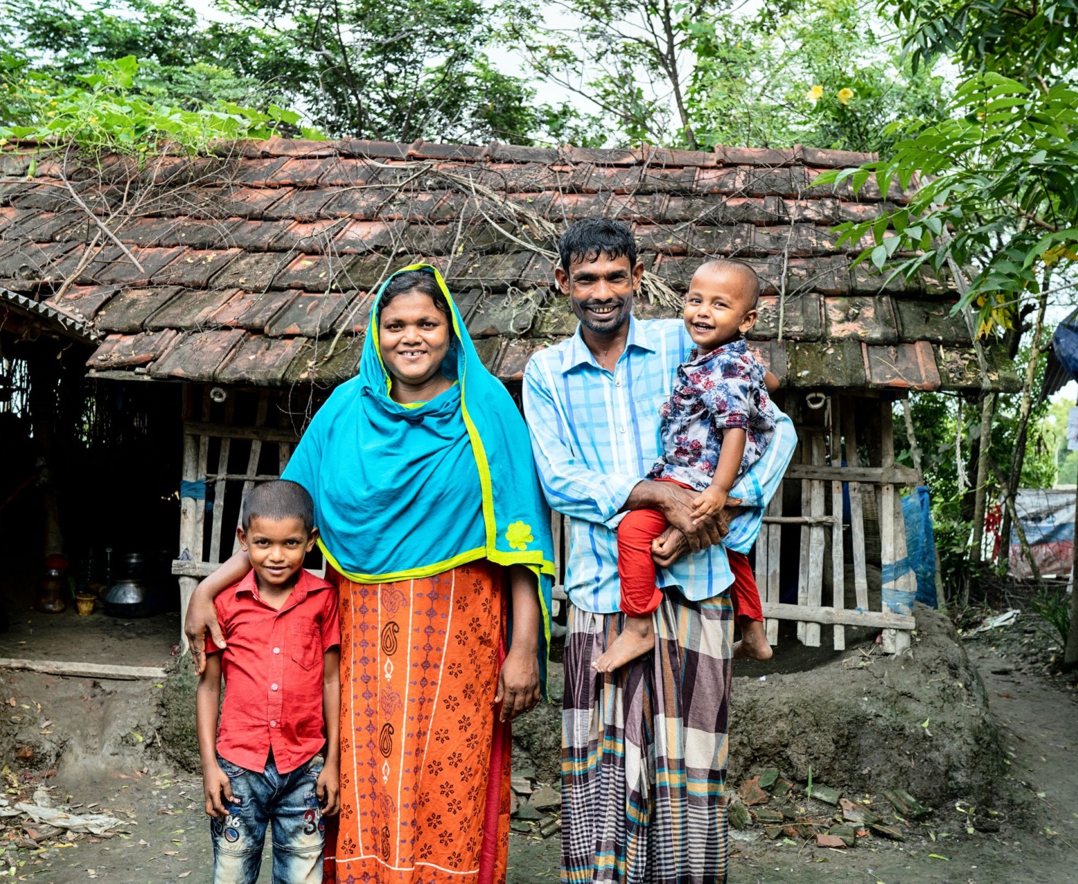A mother and a father stand smiling with their two sons outside of their home in Bangladesh.