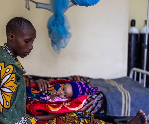 A mother sits in a hospital bed holding her baby wrapped in colorful blankets in Tanzania.